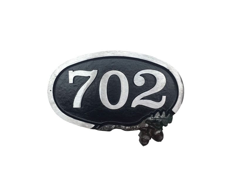 May include: A black oval-shaped house number sign with a silver border and the number 702 in white. The sign has a decorative, rustic, vine-like design at the bottom.