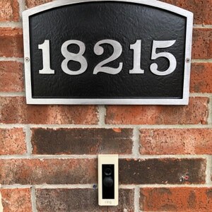 May include: A black and silver house number plaque with the number 18215 in silver lettering. A white doorbell is mounted below the plaque on a brick wall.
