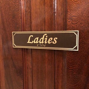 May include: A brown wooden door with a gold plaque that reads "Ladies" in gold lettering. The plaque also has braille lettering.