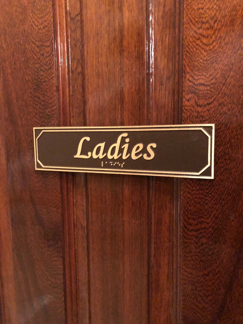 May include: A brown wooden door with a gold plaque that reads "Ladies" in gold lettering. The plaque also has braille lettering.