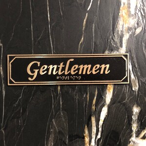 May include: A black rectangular sign with gold lettering that reads "Gentlemen" with braille below the text.