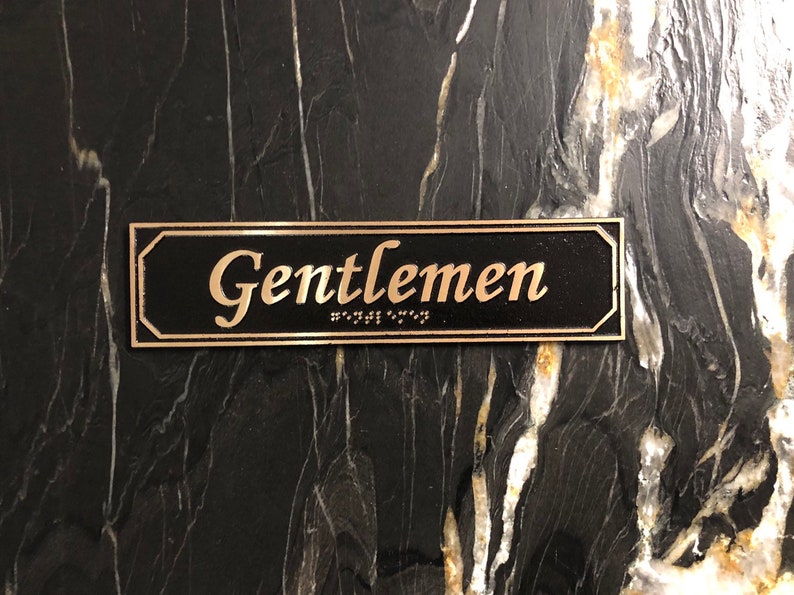 May include: A black rectangular sign with gold lettering that reads "Gentlemen" with braille below the text.