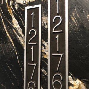 May include: Two vertical metal house number plaques with silver numbers on a dark brown background. The numbers on the left plaque are 12176 and the numbers on the right plaque are 12176.
