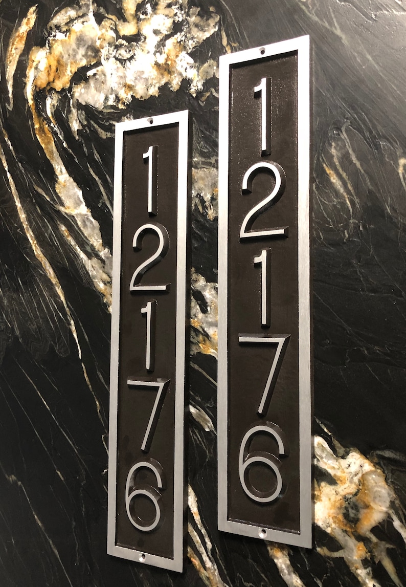 May include: Two vertical metal house number plaques with silver numbers on a dark brown background. The numbers on the left plaque are 12176 and the numbers on the right plaque are 12176.