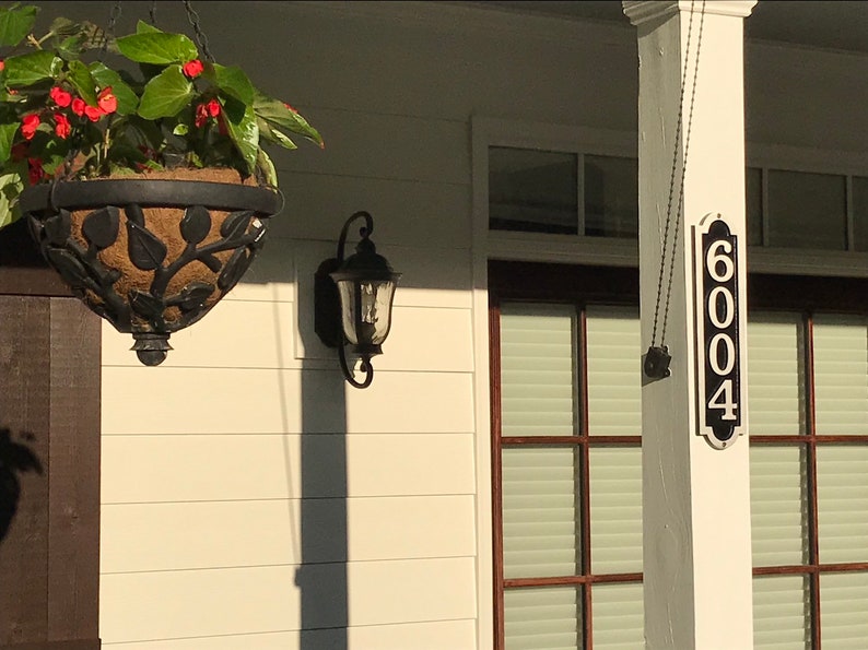 May include: A white house with a black porch and a black hanging house number sign that reads "6004". There is a black hanging light fixture on the side of the house and a hanging planter with red flowers.