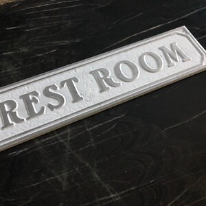 Pu&ograve; includere: Un cartello rettangolare bianco con lettere in rilievo che recita "REST ROOM" su uno sfondo nero.