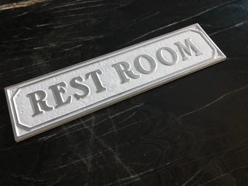 Pu&ograve; includere: Un cartello rettangolare bianco con lettere in rilievo che recita "REST ROOM" su uno sfondo nero.