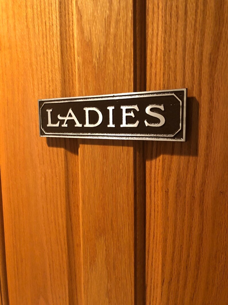 K&ouml;nnte beinhalten: Ein schwarzes und silbernes Schild mit dem Wort "LADIES" in wei&szlig;en Buchstaben.