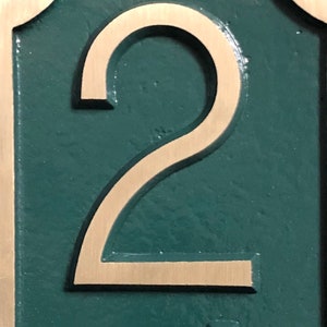 May include: A close-up of a house number plaque with the number "2" in a gold-colored font on a dark green background.