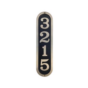 May include: A vertical, oval-shaped house number plaque with a black background and silver numbers. The numbers 3215 are displayed in a vertical line.