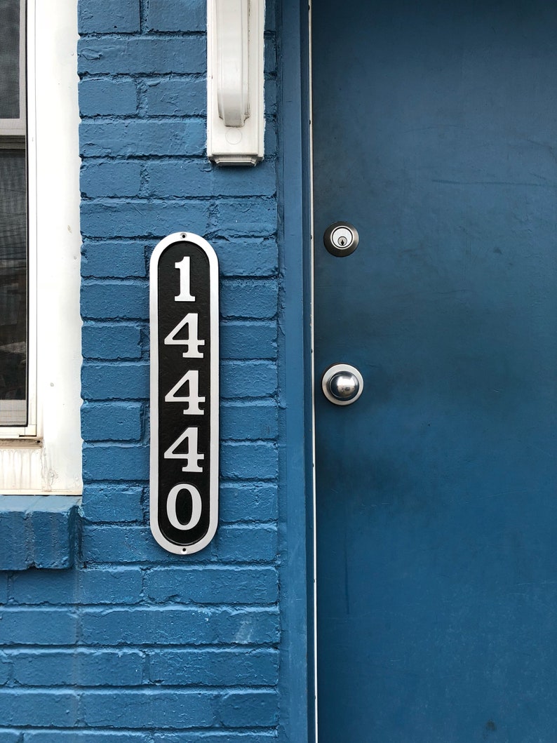 May include: A blue brick building with a blue door. The door has a silver door handle and a silver doorbell. A black and silver plaque with the number 1440 is attached to the brick wall.