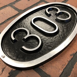 May include: A black oval-shaped house number sign with a silver border. The number 303 is embossed in the center of the sign.