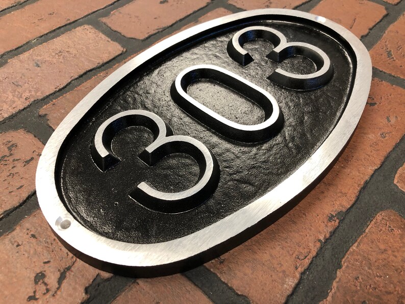 May include: A black oval-shaped house number sign with a silver border. The number 303 is embossed in the center of the sign.
