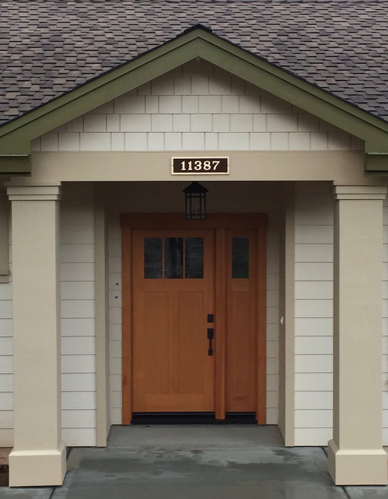 May include: A white house with a brown roof and a wooden door. The house has a green trim around the roof and a white porch with two columns. The house number is 11387.