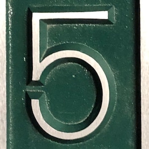 May include: A close-up of a green metal number five with a white outline. The number is raised and has a three-dimensional appearance.