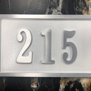 May include: A silver metal house number plaque with the number 215 embossed in white on a white background.