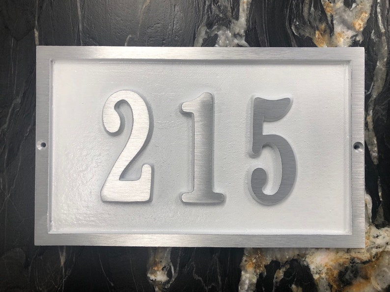 May include: A silver metal house number plaque with the number 215 embossed in white on a white background.