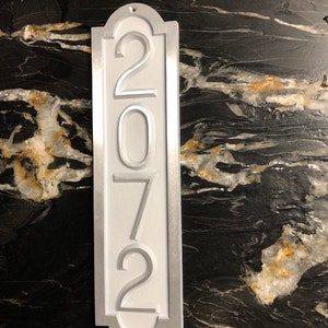 May include: A white metal house number sign with the number 2072 embossed in silver. The sign has a silver border and a rounded top.