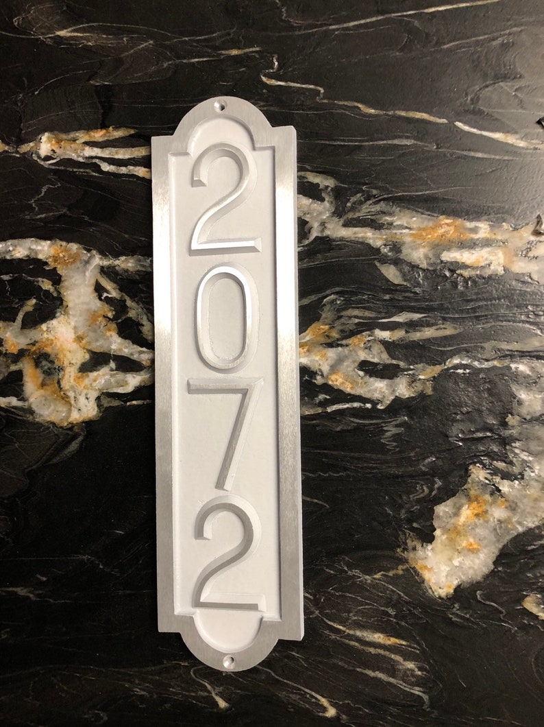May include: A white metal house number sign with the number 2072 embossed in silver. The sign has a silver border and a rounded top.