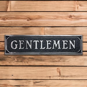 May include: A black and silver metal sign with the word "GENTLEMEN" in raised silver letters.