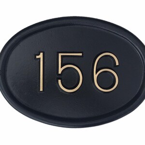 May include: Black oval-shaped house number plaque with gold-colored numbers "156" embossed on the surface.