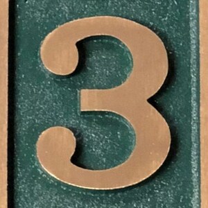 May include: A copper-coloured number three on a green background. The number is raised and has a textured surface.
