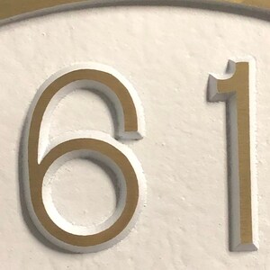 May include: Close-up of a gold-colored house number plaque with the numbers 61 embossed on a white background.