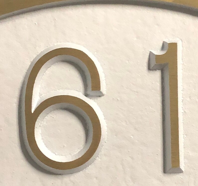 May include: Close-up of a gold-colored house number plaque with the numbers 61 embossed on a white background.