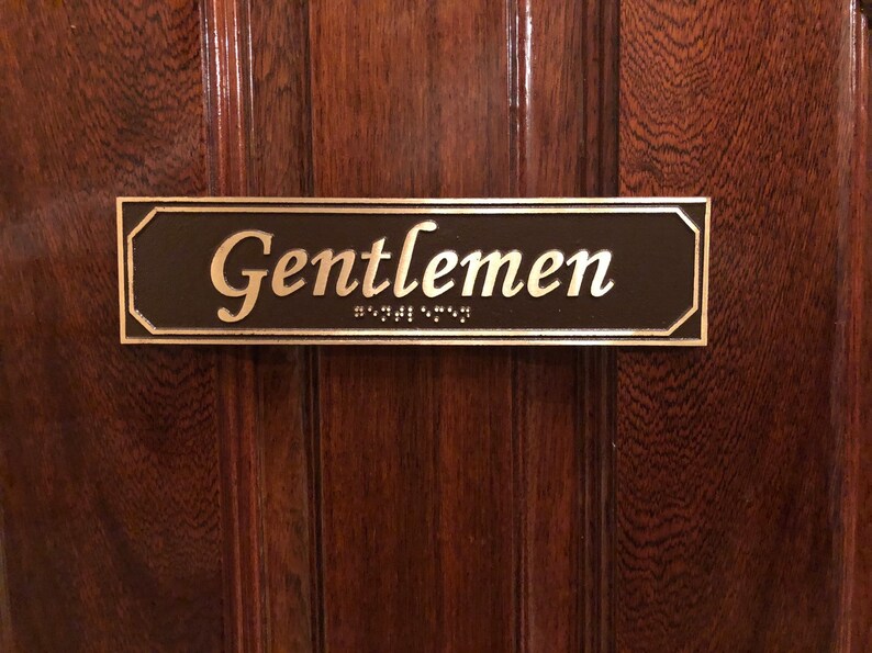 May include: A brown rectangular sign with rounded corners and a silver border. The sign reads "Gentlemen" in a gold cursive font with braille lettering below.