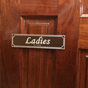 May include: A brown wooden door with a rectangular sign that reads "Ladies" in white lettering. The sign also has braille lettering.