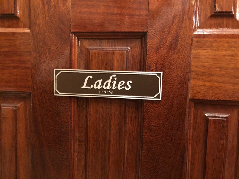 May include: A brown wooden door with a rectangular sign that reads "Ladies" in white lettering. The sign also has braille lettering.