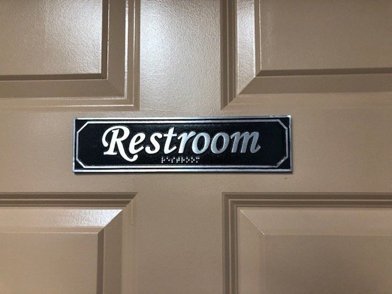 May include: A black and silver sign with the word "Restroom" in silver lettering on a beige door.