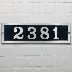 May include: Rectangular address plaque with the number 2381 in silver against a black background. The plaque has a silver border and is mounted on a white brick wall. The numbers are in a bold, serif font.