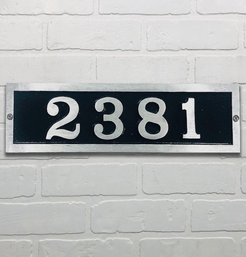 May include: Rectangular address plaque with the number 2381 in silver against a black background. The plaque has a silver border and is mounted on a white brick wall. The numbers are in a bold, serif font.