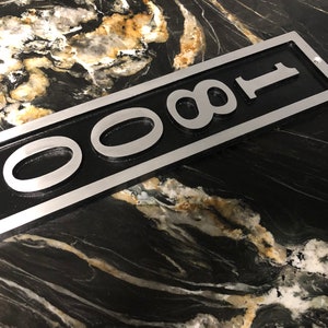 Puede incluir: Un letrero de n&uacute;mero de casa de metal plateado con fondo negro y el n&uacute;mero 0081 en letras plateadas en relieve.