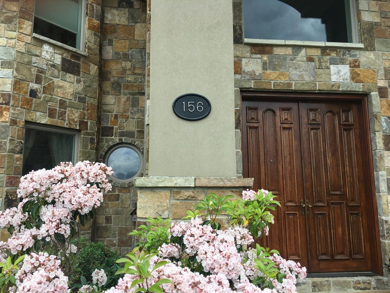 May include: A house with a stone facade and a wooden double door. The house number 156 is displayed on a black oval plaque.