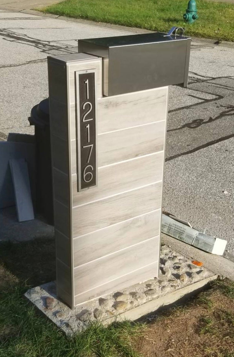 May include: A modern mailbox with a gray metal top and a white tiled base. The house number "1216" is displayed on the front of the mailbox.