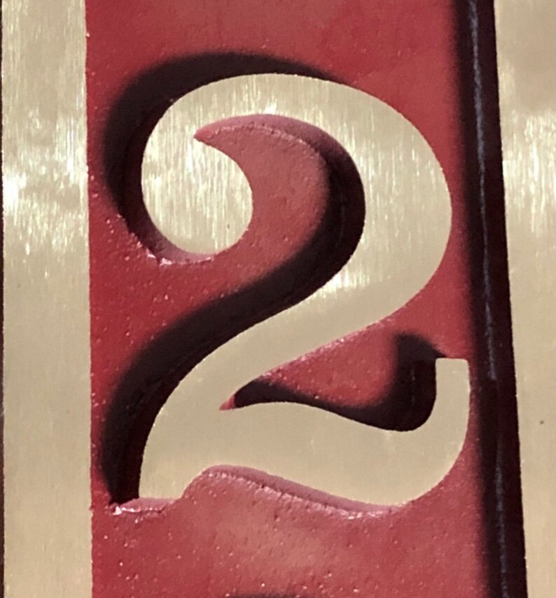 May include: A close-up of a carved wooden number two. The number is painted in a light beige colour and is set against a dark red background.