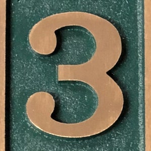 May include: A close-up of a copper-coloured number "3" on a green background. The number is raised and has a textured surface.