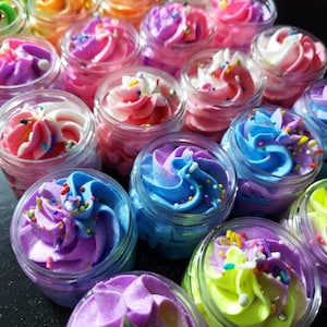 May include: A collection of small glass jars filled with colorful whipped body butter. The jars are arranged in a pyramid shape, with the largest jar at the bottom and the smallest jar at the top. The body butter is a variety of colors, including pink, purple, blue, green, and yellow. Each jar has a swirl of color and sprinkles on top.