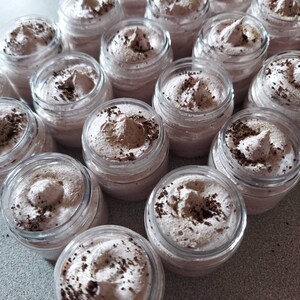 Pu&ograve; includere: Una collezione di piccoli barattoli di vetro riempiti con una sostanza di colore marrone chiaro, simile alla panna montata. I barattoli sono disposti in file su una superficie grigio chiaro. La crema ha un aspetto macchiato, probabilmente dovuto a fondi di caff&egrave; o polvere di cacao.