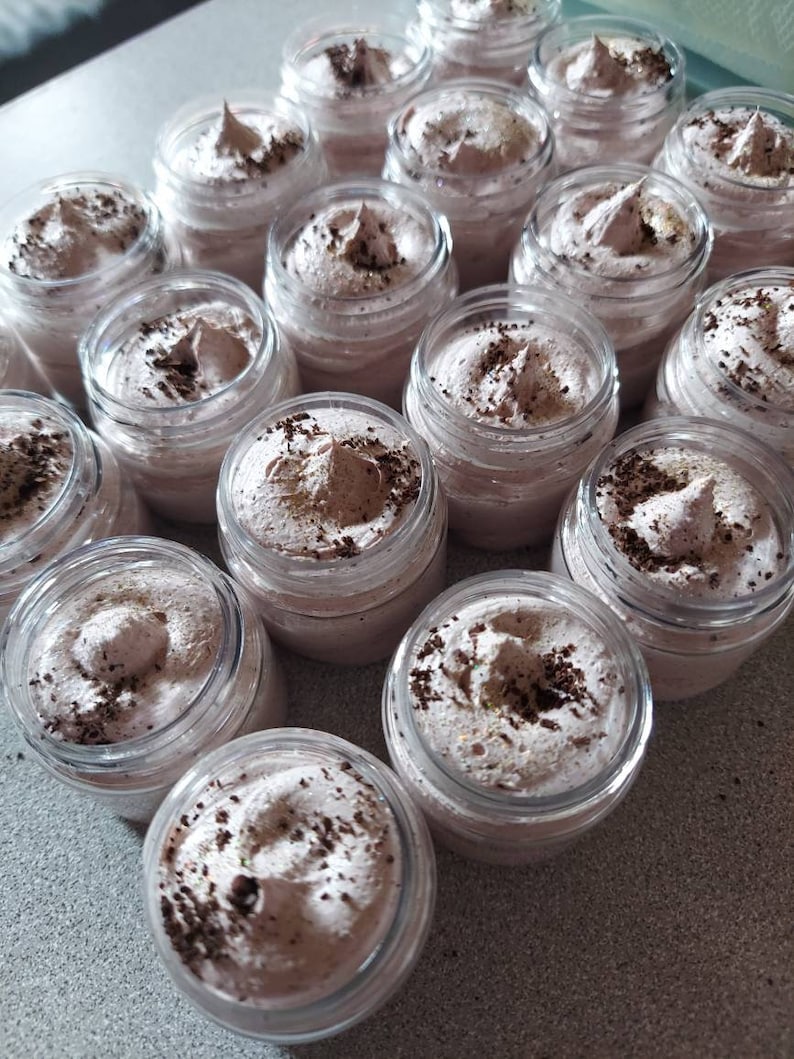 Pu&ograve; includere: Una collezione di piccoli barattoli di vetro riempiti con una sostanza di colore marrone chiaro, simile alla panna montata. I barattoli sono disposti in file su una superficie grigio chiaro. La crema ha un aspetto macchiato, probabilmente dovuto a fondi di caff&egrave; o polvere di cacao.