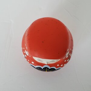May include: A red and white Japanese doll with a black and gold floral design. The doll is made of ceramic and has a round shape.