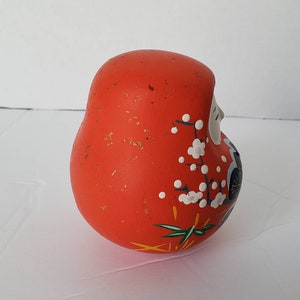 May include: A red, round, ceramic Japanese lucky charm with white and pink flower designs and gold accents. The charm is known as a "Daruma" and is a symbol of good luck and perseverance.
