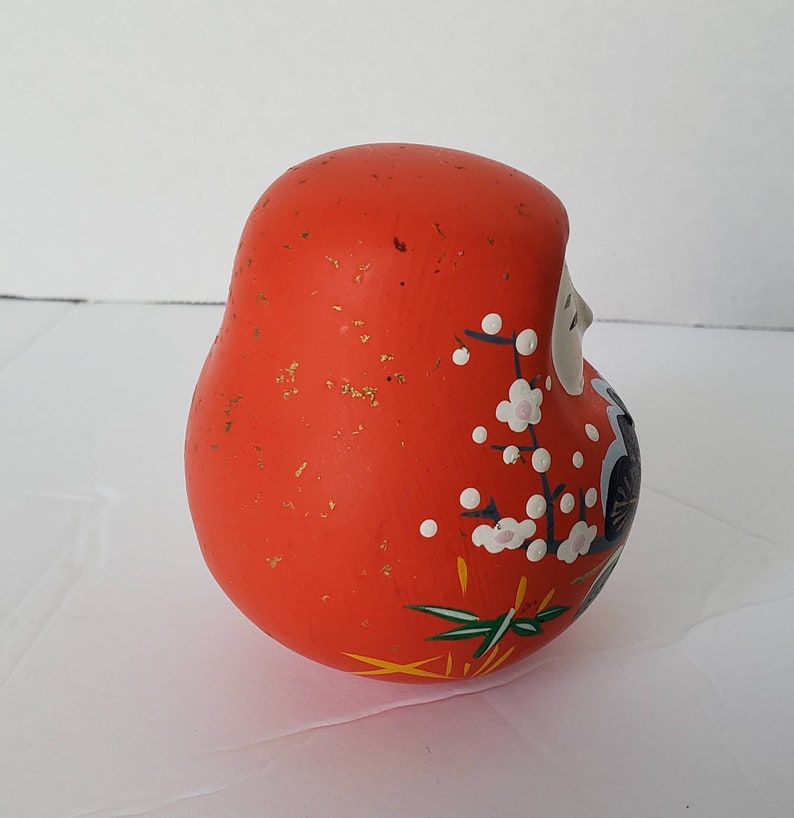 May include: A red, round, ceramic Japanese lucky charm with white and pink flower designs and gold accents. The charm is known as a "Daruma" and is a symbol of good luck and perseverance.