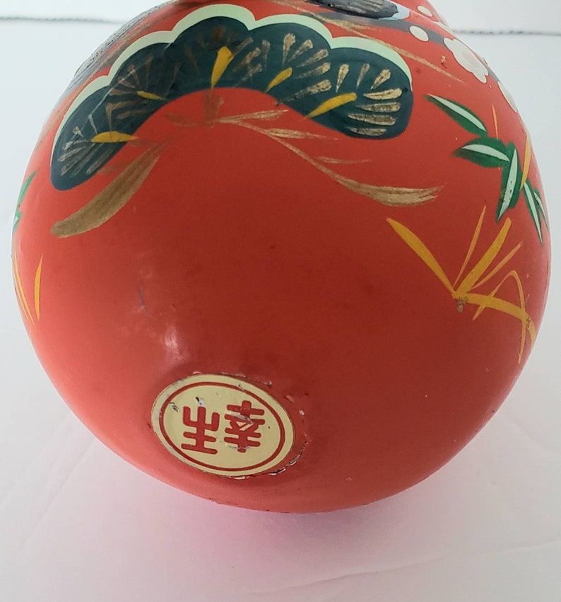 May include: A red, round, ceramic ornament with gold and green painted designs. The ornament has a white circle with the Japanese word "幸福" (Happiness) in red.