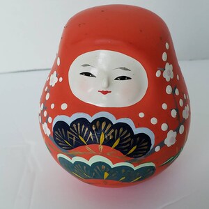 May include: A red, white, and blue Japanese traditional doll with a painted face and floral designs. The doll is round and has a slightly flattened bottom.