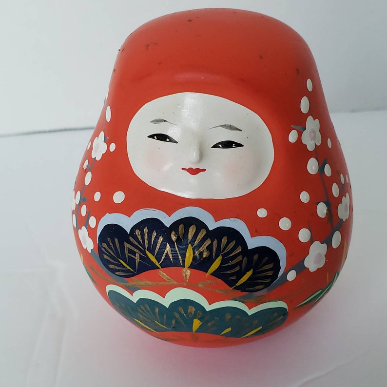 May include: A red, white, and blue Japanese traditional doll with a painted face and floral designs. The doll is round and has a slightly flattened bottom.