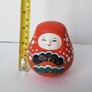 May include: A red and white Japanese Daruma doll with a painted face and floral designs. The doll is about 3 inches tall.