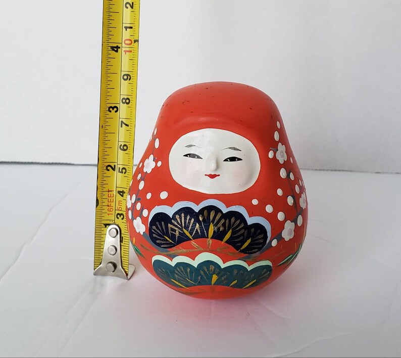 May include: A red and white Japanese Daruma doll with a painted face and floral designs. The doll is about 3 inches tall.